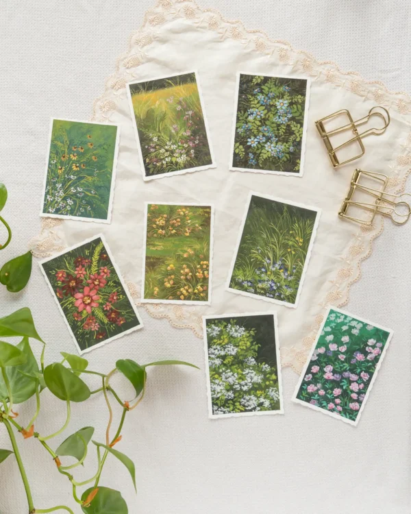 A collection of eight original postcard-sized paintings featuring various wildflowers and foliage, arranged on a lace-edged cloth with gold clips and a green plant.